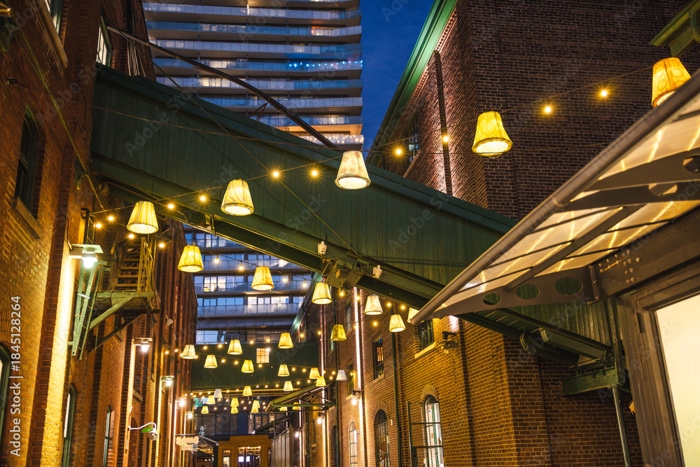 Naklejka premium Decorative hanging lamps and warm string lights illuminating a narrow brick alley in Torontos Distillery District at night.