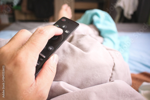 Close up of hand holding remote control while lying on bed, POV perspective of person watching TV at home.