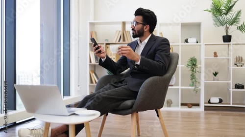An Arab businessman is seen relaxing in a modern office setting, holding a phone and drinking coffee