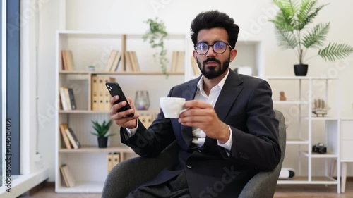 An Indian businessman in a suit smiles while holding a cup of coffee and a smartphone