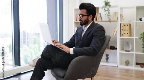 An Indian businessman wearing glasses works on his laptop while sitting in a modern office chair