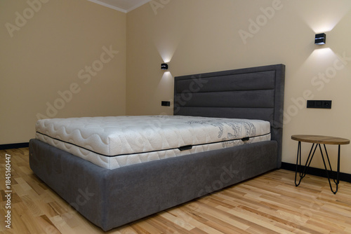 A modern bedroom interior features a double bed with a high, soft gray headboard and a light, patterned orthopedic mattress. The concept is one of comfort, sleep, furniture, and homeliness.