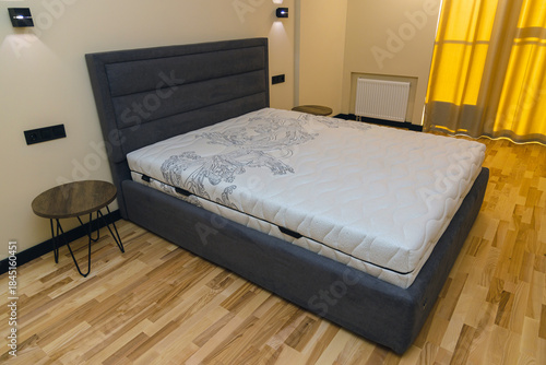 A modern bedroom interior features a double bed with a high, soft gray headboard and a light, patterned orthopedic mattress. The concept is one of comfort, sleep, furniture, and homeliness.