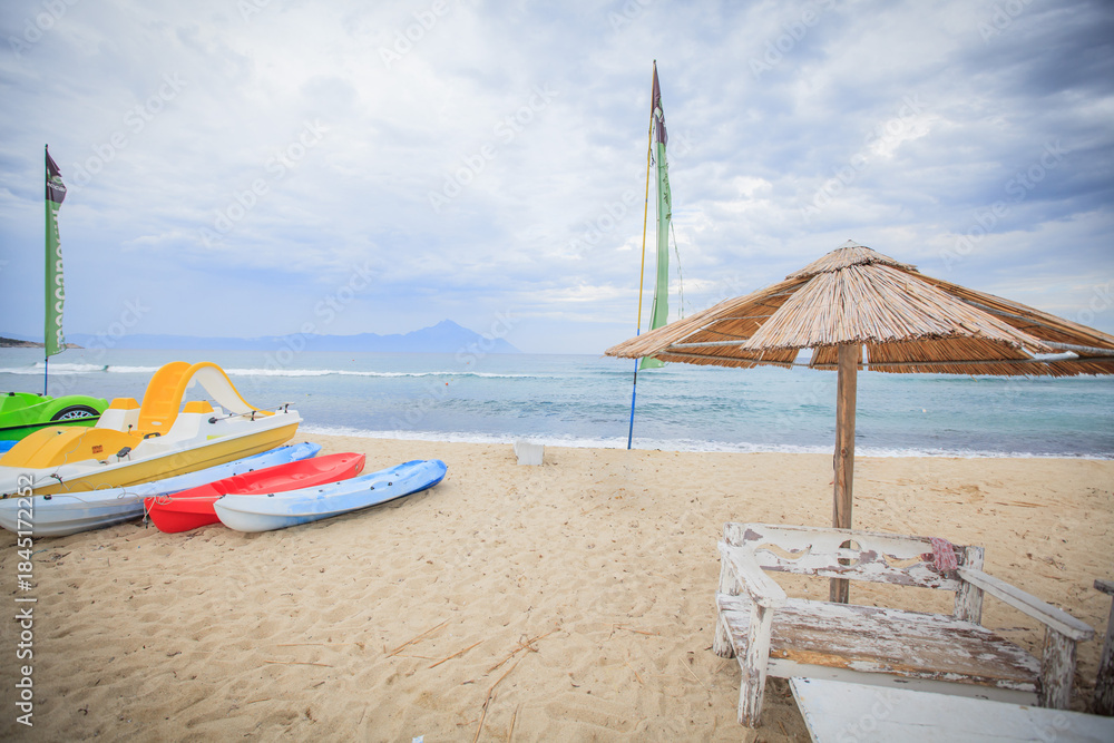 Fototapeta premium Beach scene with colorful kayaks straw umbrella and Mount Athos view, Greece