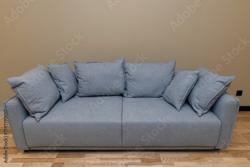 A gray fabric sofa with six large pillows sits against a solid beige wall on a wooden floor. A piece of upholstered furniture for a living room or relaxation area. Comfort and interior design concept.