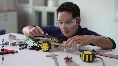 A man is working on a robot. He is wearing goggles and a blue shirt. The robot is on a table