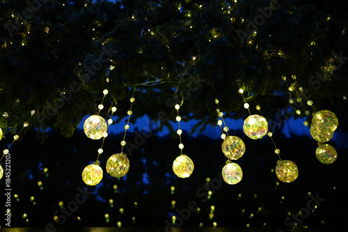 Christmas Orbs Hanging from Evergreen Branches at Night. Glowing iridescent glass ornaments and fairy lights hang from pine branches. Festive holiday bokeh background.
