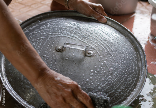 Close-up of a pair of hands scrubbing a large metal pot lid covered in soap suds and water, demonstrating housework