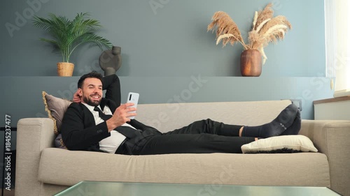 A young businessman is comfortably resting on a sofa while using his smartphone to engage in various activities, reflecting a blend of relaxation and productivity in a home setting.