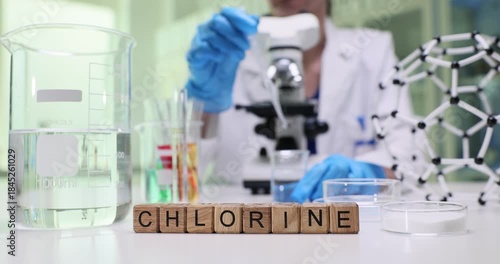 Wooden cubes on desk spell word Chlorine near clear beaker and rack. Laboratory worker pipettes liquid sample into Petri dish beside microscope