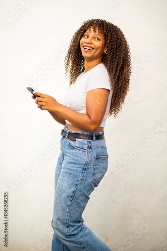 Playful young woman walking with smartphone and smiling in front of white wall
