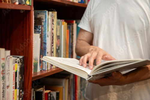 A man reading an open book while standing in front of a wooden bookshelf filled with books. Concept of education, learning, knowledge, self-study, reading habit, and intellectual.