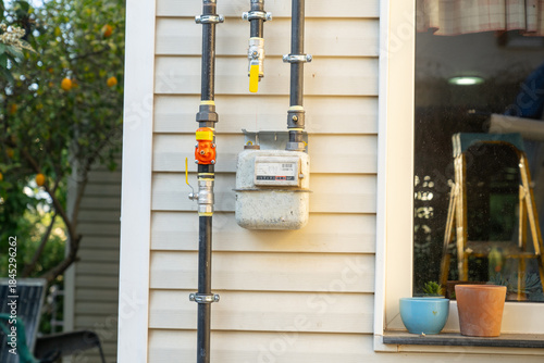 A natural gas installation and mechanical gas meter are seen mounted on the exterior house. Black metal pipes with yellow lever-operated ball valves and fittings are visible on the facade cladding.