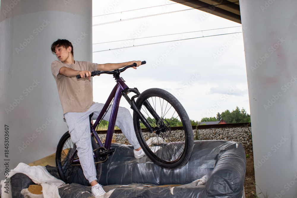 Fototapeta premium Teenage boy posing with a bicycle on an old sofa under a bridge. Urban lifestyle scene with youth, freedom and creativity in an industrial outdoor environment.