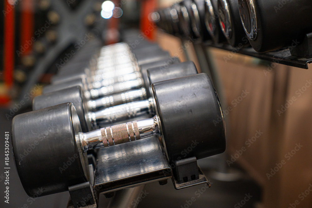 Naklejka premium Close-up of metal dumbbells neatly arranged on a rack in a modern gym. Concept of fitness, strength training, bodybuilding, workout equipment, healthy lifestyle, and exercise motivation.
