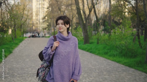 Young woman walks in a park on a sunny day wearing a purple sweater and carrying a backpack, surrounded by grassy areas and trees in the background