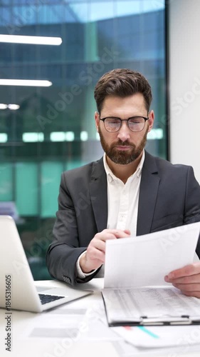 Busy businessman is doing paperwork using laptop sitting at workplace in business office. Financier in formal suit deals with financial report, checks documents, engaged in accounting. Vertical video