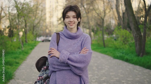 Young woman stands on a park path with trees and grass around her during a clear day in spring while smiling and crossing her arms