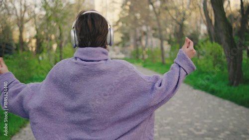 Woman dances on a paved path in a park while wearing headphones. The sun shines down and trees surround the area, creating a lively atmosphere.