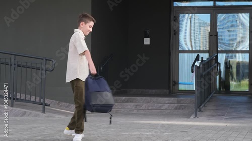 A boy is walking down a sidewalk with a backpack. He is wearing a white shirt and khaki pants