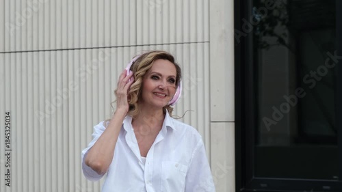 A woman wearing headphones and smiling. She is wearing a white shirt. She is walking on the sidewalk
