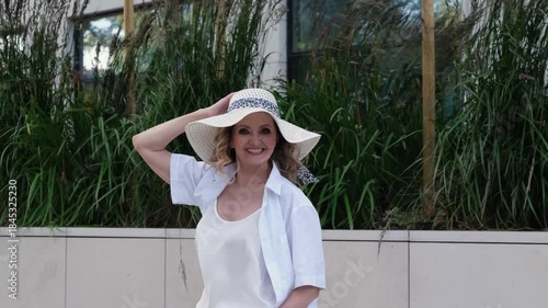 A woman wearing a white shirt and a straw hat. She is walking down the street. The hat is white and has a black and white pattern