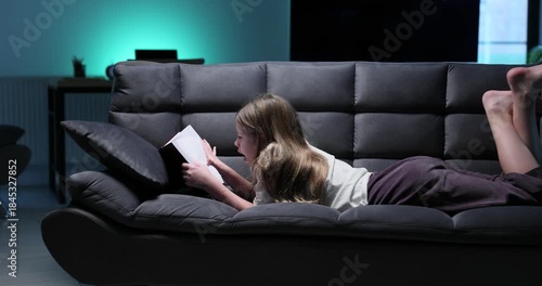 Young girl lies on dark sofa reading thick book under light. Teen girl kicks legs in air while staying absorbed in exciting adventure story pages