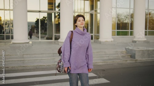 Young woman walks across a crosswalk near a building with columns during the late afternoon, carrying a bag on her shoulder and smiling softly