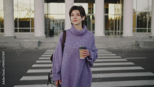 Woman stands at a crosswalk in front of a large building holding a cup and wearing a sweater during the late afternoon sun