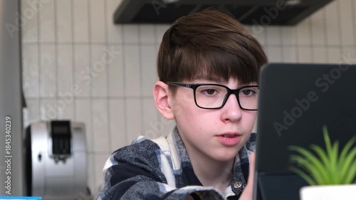 A boy wearing glasses is looking at a laptop. A potted plant is next to him. A coffee maker is also visible in the background
