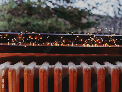 Photo of a window with a view of the forest, a windowsill with a Christmas garland and a retro cast iron radiator heater