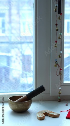 Close-up vertical shot of a brass singing bowl and mallet on a windowsill next to a decorative string of small brass bells and beads, set against a bright, blurred window showing an urban background