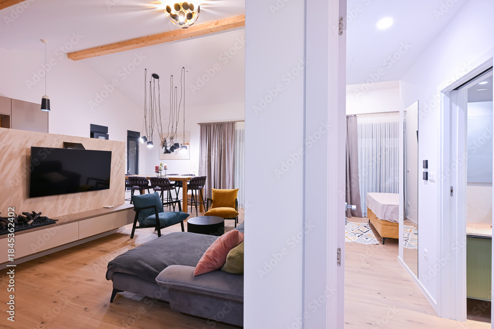 Obraz premium Modern open-plan living and dining area with an integrated kitchen, featuring a comfortable sofa, stylish armchairs, and a bar-height dining table. Wooden floors and ceiling beams enhance this