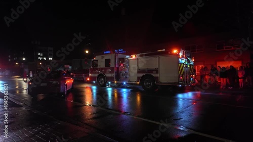 Langford, BC, Canada. December 15, 2025: A fire truck with flashing lights responds on a wet city street at night, reflecting urgency and safety, as a crowd gathers in the background.