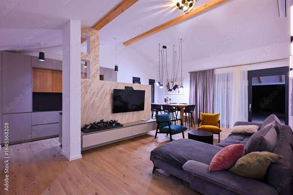 Obraz premium Modern open-plan living and dining area with an integrated kitchen, featuring a comfortable sofa, stylish armchairs, and a bar-height dining table. Wooden floors and ceiling beams enhance this