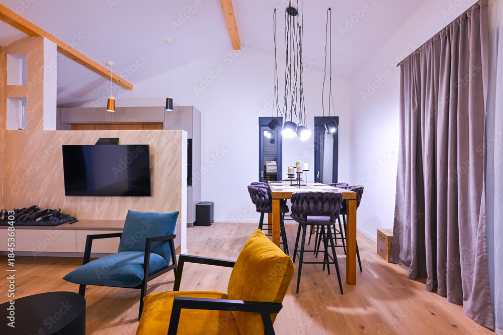 Obraz premium Modern open-plan living and dining area with an integrated kitchen, featuring a comfortable sofa, stylish armchairs, and a bar-height dining table. Wooden floors and ceiling beams enhance this