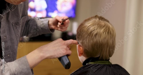 Mother cutting hair to child with comb and clippers while boy watching cartoons at home