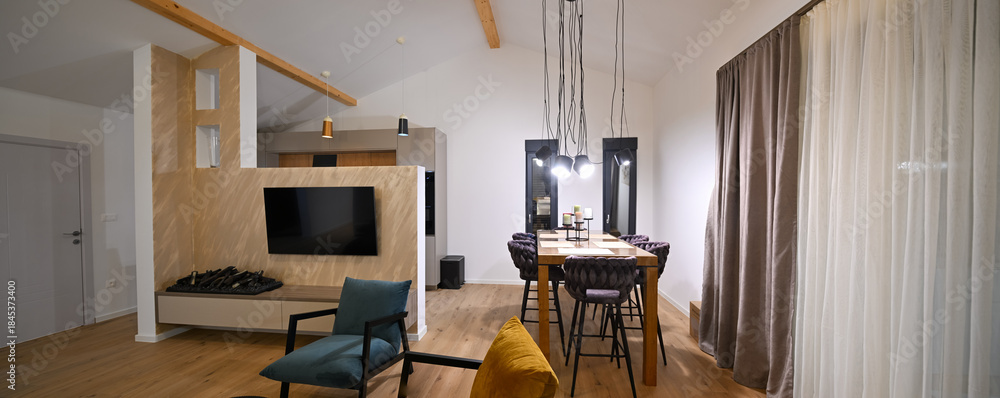 Obraz premium Modern open-plan living and dining area with an integrated kitchen, featuring a comfortable sofa, stylish armchairs, and a bar-height dining table. Wooden floors and ceiling beams enhance this