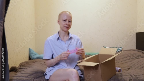 Bald woman after chemotherapy sitting on the bed and opening a cardboard box, concept of recovery, new beginnings, and peaceful daily life at home