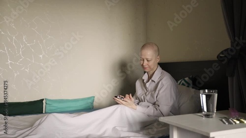 Young bald woman with cancer using mobile phone in bedroom, lying in bed under white blanket, chatting with family and friends, reading messages and social media, emotional support during chemotherapy