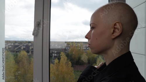 Bald woman after cancer treatment standing by the window, looking outside with thoughtful expression. The autumn landscape outside contrasts with her calm, 
