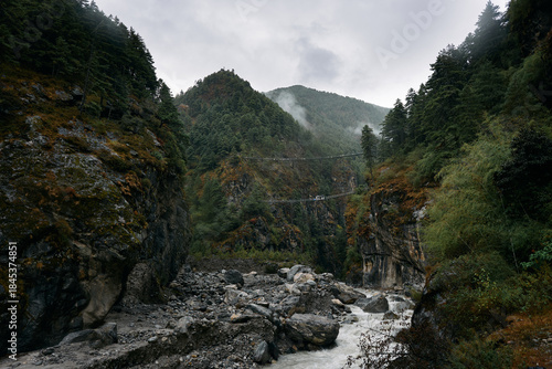 A bridge connects two sides of a valley above a river. The scene shows tall mountains and trees on either side. Clouds cover the sky and there is water flowing swiftly below