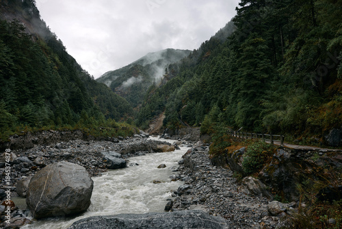 A river moves swiftly through a rocky terrain with large stones and trees on both sides. Mist hangs over the mountains in the distance. The day is cloudy and gray