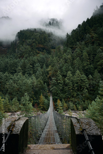 A long suspension bridge stretches across a green valley. Thick forests surround the area while low clouds cover the mountains. The scene captures nature's quiet beauty and vastness