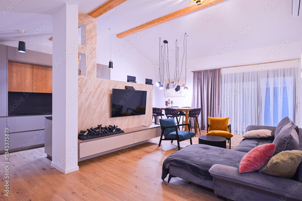 Obraz premium Modern open-plan living and dining area with an integrated kitchen, featuring a comfortable sofa, stylish armchairs, and a bar-height dining table. Wooden floors and ceiling beams enhance this