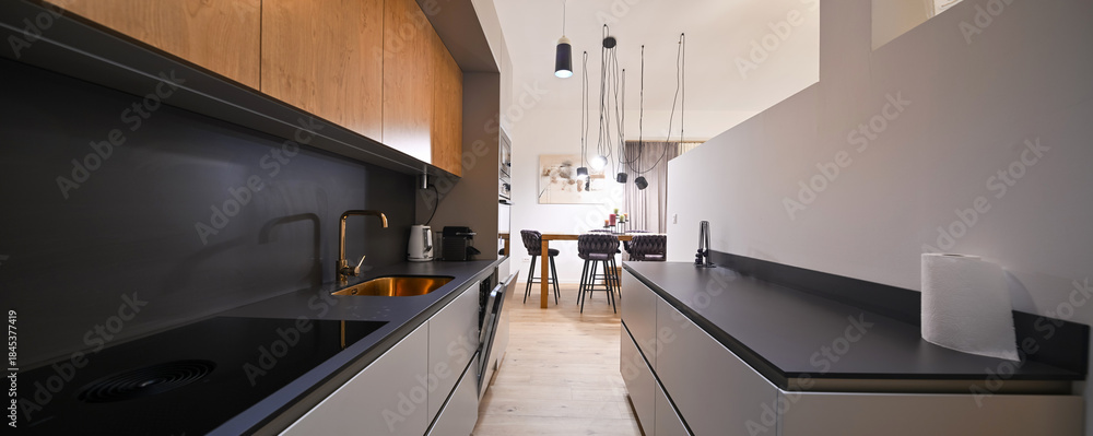 Obraz premium Modern open-plan living and dining area with an integrated kitchen, featuring a comfortable sofa, stylish armchairs, and a bar-height dining table. Wooden floors and ceiling beams enhance this