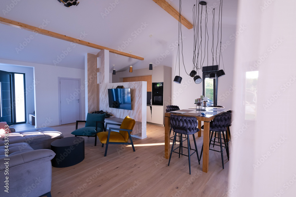 Obraz premium Modern open-plan living and dining area with an integrated kitchen, featuring a comfortable sofa, stylish armchairs, and a bar-height dining table. Wooden floors and ceiling beams enhance this