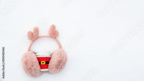 Cup of drink in knitted clothes of Santa Claus and fur pink winter earmuffs on snow background