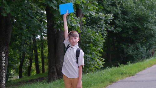 Cute schoolboy in schoolyard with school supplies holding book up, back to school concept