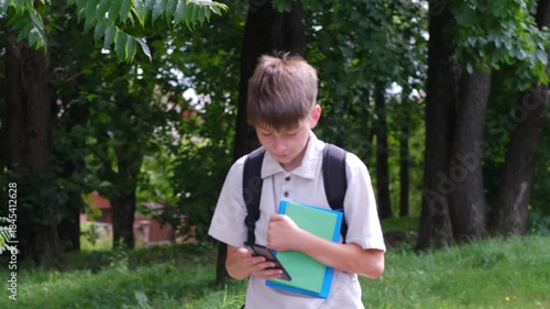 A schoolboy boy with a phone in his hand and books walks down the street